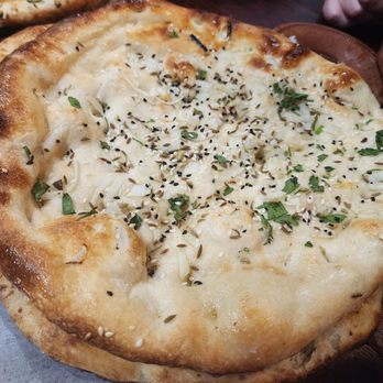 Clay Handi - Garlic and ginger Naan - A photograph features a naan bread topped with chopped herbs, black sesame seeds, and other seasonings. The bread has a golden-brown crust with slightly charred spots and is served on a plate. In the background, part of another dish and a person's arm are visible, indicating a dining setting. - Halal Near Me - Buffalo, New York