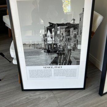 AlphaGraphics - The image shows a framed black-and-white photograph of Venice, Italy, placed on a wooden floor and leaning against a wall. The photograph depicts buildings along a canal with chairs and water in the foreground. The frame is black, and the photograph includes a caption with the title "Venice, Italy." The reflection of people and a window can be seen on the glass of the frame. - Printing Services Near Me - Vista, California
