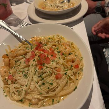 Bonefish Grill - A white bowl contains shrimp pasta with a creamy sauce, topped with chopped tomatoes and parsley, and sprinkled with grated cheese. The dish is placed on a white tablecloth, with a fork resting inside the bowl. In the background, there is another bowl of soup with two spoons, and part of a person's arm and hand are visible, indicating a dining setting. - Seafood Near Me - Brookfield, Wisconsin