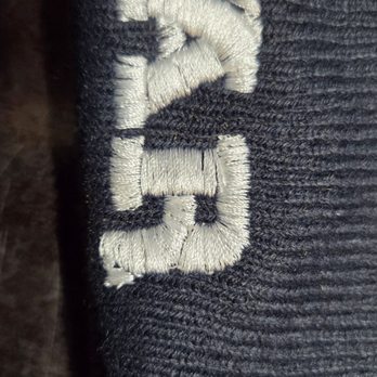 Icustomize - Letter R is tilting down and badly done. - The image shows a close-up of black fabric with white embroidered text. The embroidery appears slightly frayed or uneven, and the fabric has a textured, ribbed surface. The background is dark and indistinct, emphasizing the embroidered detail. - Screen Printing/T-Shirt Printing Near Me - Montebello, California