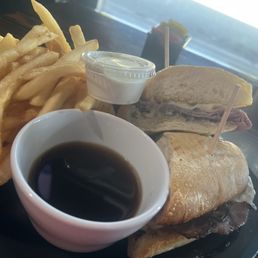 French Dip with Swiss Cheese - The image shows a French dip sandwich with Swiss cheese on a toasted bun, with slices of roast beef inside. It is served with a side of French fries, a small cup of dipping sauce, and a cup of black coffee. The meal is presented on a dark plate, with a background indicating a casual dining setting.