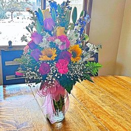 A floral arrangement featuring roses, sunflowers, lilies, and other flowers in a glass vase on a wooden table by a window.