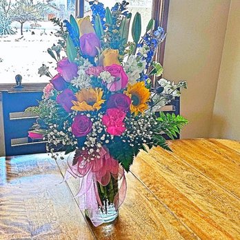 Vail Flowers - A floral arrangement featuring roses, sunflowers, lilies, and other flowers in a glass vase on a wooden table by a window. - Florists Near Me - Vail, Arizona