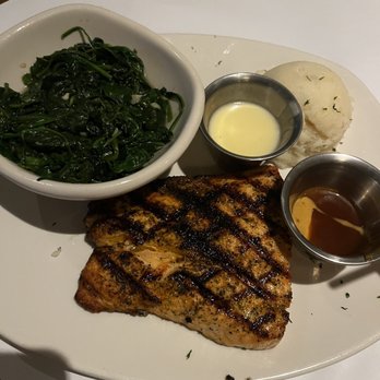 Bonefish Grill - Atlantic Salmon* - The image shows a white plate with a grilled salmon fillet that has visible grill marks and a seasoned crust. Accompanying the salmon are a serving of mashed potatoes, a bowl of sautéed greens, and small metal containers of butter and sauce. The presentation is on a white surface, likely in a restaurant setting. - Seafood Near Me - Gambrills, Maryland