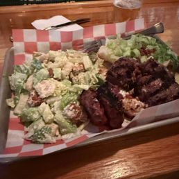 Steak Tips - A tray holds a Caesar salad and grilled meat, with a beer glass and utensils on a bar counter, suggesting a casual dining setting.