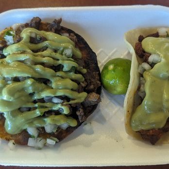Tacos Tijuana - Carne asada vampiro Al pastor taco - Two tacos filled with grilled meat, topped with chopped onions and drizzled with green sauce, served on a white foam tray. A lime is placed between the tacos, and the background is a wooden surface. - Tacos Near Me - Mesa, Arizona