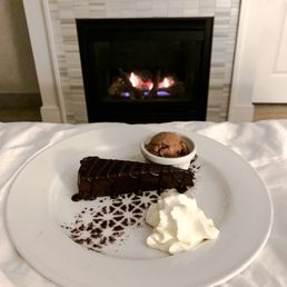 A white plate holds a slice of chocolate cake with chocolate drizzle, a dollop of whipped cream, and a small bowl of ice cream. The plate is positioned on a bed or soft surface, with a fireplace burning in the background, creating a cozy setting.