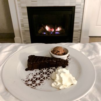 Restaurant 301 - A white plate holds a slice of chocolate cake with chocolate drizzle, a dollop of whipped cream, and a small bowl of ice cream. The plate is positioned on a bed or soft surface, with a fireplace burning in the background, creating a cozy setting. - Breakfast & Brunch Near Me - Duluth, Minnesota