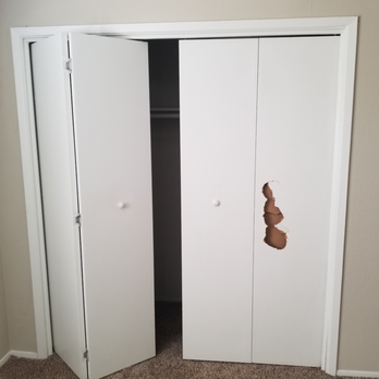 Knotty's Custom Carpentry - A closet with white sliding doors is shown, with one door partially open and the other closed. The right door has a large, irregular hole with torn edges, exposing the closet interior. The closet is set into a beige wall, and the floor is carpeted in brown. - Handyman Near Me