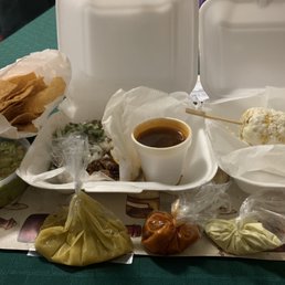 From Left to Right: Guacamole with chips, Birria QuesaTaco, consomé and elote. At the front, different small baggies of hot sauces. From Left to Right: Guacamole with chips, Birria QuesaTaco, consomé and elote. At the front, different small baggies of hot sauces.
