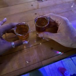 Two people are holding shot glasses filled with a dark-colored beverage, possibly alcohol, over a wooden bar or table. One person is wearing a ring on their finger, and the other has a visible hand with no jewelry. The background shows keys, a wallet, and some electronic devices on the wooden surface, indicating a casual social setting.
