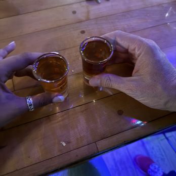Western Bar & Cafe - Two people are holding shot glasses filled with a dark-colored beverage, possibly alcohol, over a wooden bar or table. One person is wearing a ring on their finger, and the other has a visible hand with no jewelry. The background shows keys, a wallet, and some electronic devices on the wooden surface, indicating a casual social setting. - American Near Me - Cloudcroft, New Mexico