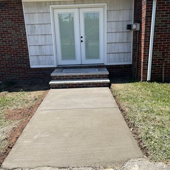 Pro Touch Roofing  - Brand new steps and walkway =] - The image shows a newly constructed concrete walkway leading to a small brick staircase with two steps, which provides access to a white double door with glass panels. The house exterior features a combination of brick and siding, with a gutter and downspout visible on the right side. The area around the walkway has some patches of grass and dirt, indicating recent landscaping or construction activity. - Roofing Near Me