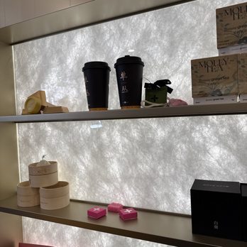 Molly Tea - A retail display shelf features various tea-related items, including black takeaway cups, round wooden boxes, pink boxes, and a black box, arranged on two levels. The background is a textured, illuminated surface that appears to be frosted or patterned glass, providing a diffused light behind the products. The scene suggests a tea shop or specialty tea store setting. - Bubble Tea Near Me - Brooklyn, New York