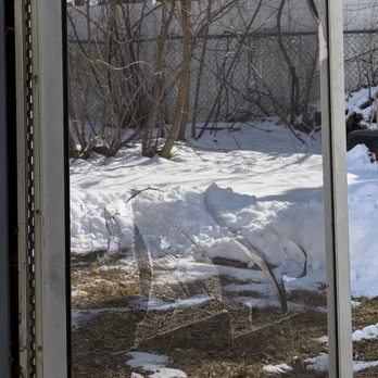 Safe Responsible Movers - The image depicts a glass door with a metal frame, looking out onto a winter landscape. Outside, there is snow covering the ground, with some patches of dirt and debris visible near the door. Leafless trees and a chain-link fence are seen in the background, indicating a cold, winter setting. The scene is illuminated by natural daylight, and the door's reflection shows part of the interior space. - Movers Near Me - Boston, Massachusetts