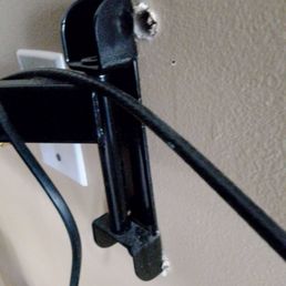 This is how David (The owner) left my TV mount. - A black TV wall mount is partially detached from the wall, with torn drywall and exposed wall material around it.