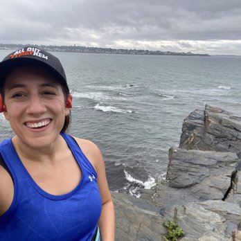Cliff Walk - A woman wearing a blue athletic tank top and a black cap is taking a selfie at a rocky shoreline. The background features water with small waves, a distant landmass, and a cloudy sky. She is smiling and appears to be outdoors near a body of water, possibly during a walk or run along the coast. - Parks Near Me - Newport, Rhode Island
