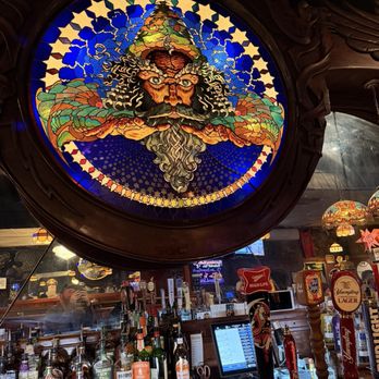 Stober's - A stained-glass window with vibrant colors shows a wizard with a pointed hat, flowing beard, and intense eyes. The window is framed by dark wood and is positioned above a bar area with bottles of alcohol, beer taps, and a touchscreen register. The setting appears to be inside a bar or pub with decorative lighting and a mirror reflecting part of the interior. - Bars Near Me - Lansing, Michigan