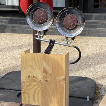 Stable DC - Heaters! - Two vintage-style outdoor heaters with metal grates and protective cages are mounted on a horizontal metal pipe, which is attached to a wooden block. The heaters are positioned in front of a building with black brick steps and large windows. The scene appears to be set outside a restaurant or cafe, with a red curtain or fabric hanging behind the heaters. The ground is paved with small stones or gravel. - Modern European Near Me - Washington, DC, District of Columbia