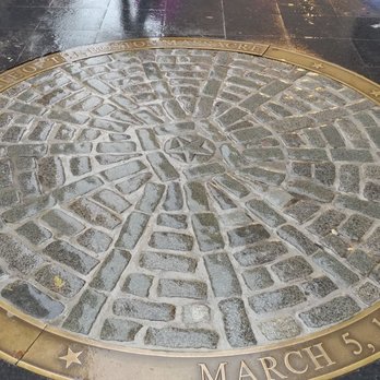 Boston Massacre Memorial - The image shows a circular plaque embedded in the pavement, made of stone and metal. The plaque features a cobblestone pattern in the center, surrounded by a metal border with inscriptions. The inscriptions include the phrase "Site of the Boston Massacre" and the date "March 5, 1770." The surrounding pavement is dark and wet, indicating recent rain or cleaning. - Landmarks & Historical Buildings Near Me - Boston, Massachusetts