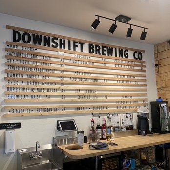 Downshift Brewing: Outpost 1706 - The image shows the interior of Downshift Brewing Co, with a large wooden menu board on the wall listing beers and prices. The bar area includes bottles of alcohol, a coffee machine, a small sink, and a tablet device. The decor features a mix of white and wooden walls, with a ceiling-mounted track lighting system. The setup suggests a casual brewery and cocktail bar environment. - Beer, Wine & Spirits Near Me - Albuquerque, New Mexico