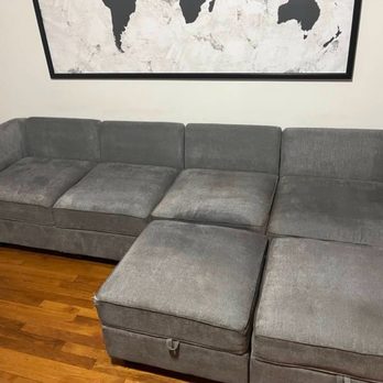 Beeclean - After (still wet) - A gray fabric sectional sofa with multiple cushions and a chaise lounge section is situated against a wall in a room with wooden flooring. The sofa appears used, with some visible wear on the cushions. A large world map artwork hangs on the wall above the sofa, and part of a shelving unit is visible on the left side of the image. The room has a neutral color palette and a modern decor style. - Carpet Cleaning Near Me