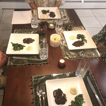 Chef Devlin O'Connor - The image shows a rectangular wooden dining table set for three people, with three white square plates containing food, including dark sauce, broccoli, and a dollop of mashed potatoes. There are glasses of water and wine, candles in the center, and green placemats with tassels. The setting appears to be in a restaurant or dining area with tiled flooring and a neutral wall background. - Personal Chefs Near Me