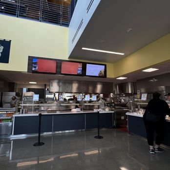 Latitude - Overview of the Hawaiian Grill station - The image shows the interior of a cafeteria or food court with a high ceiling and modern design. The serving counter is equipped with stainless steel kitchen appliances and a glass display case. Above the counter, three digital menu screens are visible, displaying menu options. A person is standing at the counter, possibly placing an order. The floor is polished concrete, and the ceiling has wooden beams and hanging lights, contributing to a contemporary atmosphere. - Middle Eastern Near Me - Davis, California