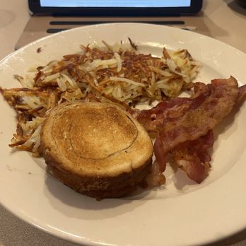 Ned's Diner - That's the cinnamon toast. Made with cinnamon bread! - A breakfast meal on a white plate includes shredded hash browns, a golden-brown biscuit, and two strips of cooked bacon. The plate is placed on a table with a tablet or menu in the background, indicating a dining setting. - Breakfast & Brunch Near Me - Belvidere, Illinois