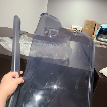 Affordable Moving Solutions - A person is holding a large, transparent, cracked glass panel or windshield with visible damage and scratches. The background shows a storage area with boxes, furniture, and a pet carrier, indicating an indoor warehouse or storage facility. - Movers Near Me - Bridgeton, Missouri