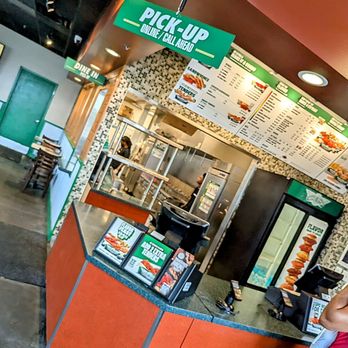 Wingstop - Anyone working here willing to come take an order? Of course not. - Counter area of Wingstop restaurant with a menu board overhead and a pick-up sign, showing the kitchen behind and a customer at the counter. - Chicken Wings Near Me - Forestville, Maryland