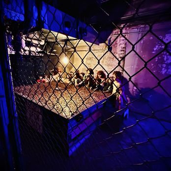 Hanny's - Creepy dolls in the basement - People are seated at a long table in a dark, industrial-style space with purple and yellow lighting, viewed through a chain-link fence. - Lounges Near Me - Phoenix, Arizona