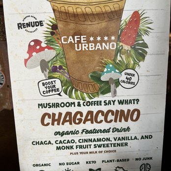 Cafe Urbano - Chagaccino poster on the counter showing everything in this tasty drink (I don't think it's listed on their regular menu) - The menu sign from Cafe Urbano features a large illustration of a coffee cup labeled 'Chagaccino,' surrounded by colorful mushroom and tropical leaf illustrations. The sign highlights the drink as an organic featured beverage with flavors like chaga, cacao, cinnamon, vanilla, and monk fruit sweetener. It emphasizes health benefits such as stress relief, immune boost, anti-aging, endurance, and cognitive function, and mentions options like organic, no sugar, keto, plant-based, and no junk. Additional notes include 'boost your coffee' and 'under 40 calories.' - Cafes Near Me - Chicago, Illinois