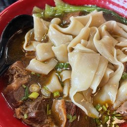 Hand Pulled Noodles - A bowl of beef noodle soup contains hand-pulled noodles, beef chunks, green vegetables, and chopped green onions in a broth. The noodles are wide and slightly curled, and the beef appears tender. The broth is dark and flavorful, with visible oil and seasoning. The bowl is red, and a black spoon rests inside. The background shows a table with other items, indicating a restaurant setting.