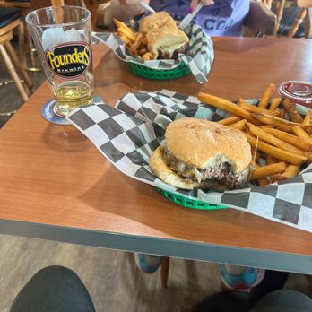 Papa Jake's - Mushroom Swiss burger and my daughters meatball sub yumm - The image shows two burgers with melted cheese and beef patties, served with a side of French fries in green baskets lined with black-and-white checkered paper. A glass of beer with a Founders Brewing logo is on the table, along with a small container of ketchup. A person wearing a purple T-shirt is seen in the background, cutting into one of the burgers. The setting appears to be a casual dining restaurant with wooden tables and chairs. - Sports Bars Near Me - Teaticket, Massachusetts