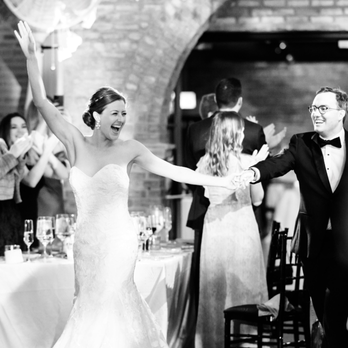 Big City Bride - A bride in a strapless wedding gown and a groom in a tuxedo are dancing together at their wedding reception. The bride has her arm raised and is smiling broadly, while the groom holds her hand and smiles back. Guests in formal attire are standing around, clapping and celebrating. The setting appears to be an indoor venue with brick walls and a table with glasses and tableware in the background. - Party & Event Planning Near Me - Chicago, Illinois