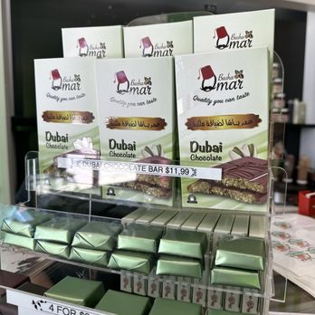 Ranoush - The image shows a retail stand with Dubai chocolate bars in green packaging, priced at $11.99. Below, there are wrapped chocolates in green foil, with a sign indicating 4 for $9.99 and individual prices of $2.99. The display is organized with clear pricing labels, and the background includes a blurred view of a store interior with various items and signage. - Waffles Near Me - Lexington, Kentucky