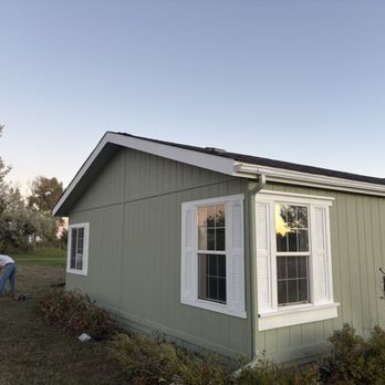 Durango Colorscapes - A person is working outside a light green house with white window shutters, planting a tree in the yard during daylight. - Painters Near Me