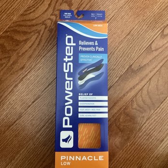 The Running Store - Power step Insole - The image features a tall, rectangular box of PowerStep Pinnacle Low insoles resting on a wooden floor. The box is primarily blue and orange, with product branding, features, and benefits printed on it, including relief for plantar fasciitis, overpronation, foot, arch, heel pain, and sore, aching feet. The packaging displays an illustration of the insoles and emphasizes their proven clinical effectiveness. - Sports Wear Near Me - Gainesville, Virginia