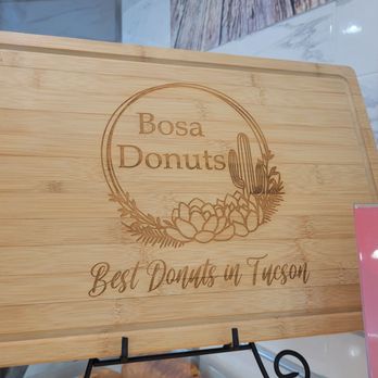 BoSa Donuts - A rectangular wooden tray features the Bosa Donuts logo, which includes a circular design with the business name and an illustration of a cactus and succulents. Below the logo, the phrase 'Best Donuts in Tucson' is engraved. The tray is placed on a display stand inside a store, with part of a pink sign and a marble surface visible in the background. - Donuts Near Me - Tucson, Arizona