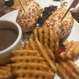 Ribs - The image shows a plate with two cheeseburgers, each with toasted buns topped with melted cheese and skewers. The burgers are served with a side of waffle fries and a small cup of dipping sauce. The setting appears to be a casual dining restaurant, with other dishes and condiments visible in the background.