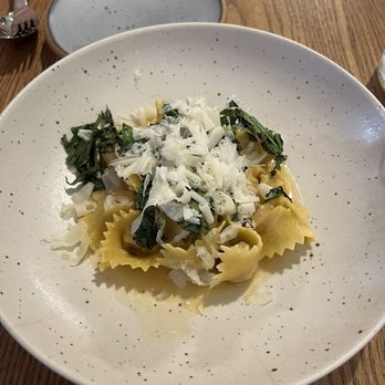 Wildweed - Chicken Ranger tortelli - A dish of pasta with shredded cheese and greens is served on a speckled ceramic plate on a wooden table. - New American Near Me - Cincinnati, Ohio
