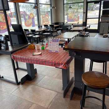 Taco Bell - Decorative table with information on becoming an employee. - A table with a red plaid tablecloth is decorated with small holiday-themed items, including a snowman figure, a Christmas tree, and a Santa figure. An informational flyer is also placed on the table. The restaurant interior features multiple tables and chairs, large windows with promotional images, and a counter area with a cash register. The space is well-lit with natural light and has a tiled floor. - Tacos Near Me - Cuba, Missouri