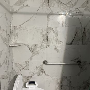 P G Builders - Shower - The image shows a bathroom with walls covered in large marble-patterned tiles in shades of white and gray. A white toilet with a black handle is positioned in the lower part of the image. Metal grab bars are installed on the walls, indicating accessibility features. A small soap dish is mounted on the wall near the corner. The lighting creates a reflection on the tiles, and part of a colorful shower curtain is visible on the right side. - General Contractors Near Me - Bethesda, Maryland