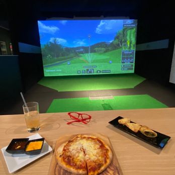 X-Golf - Westbury - a pizza on a table with a tv in the background - Golf Lessons Near Me - Westbury, New York X-Golf - Westbury - a pizza on a table with a tv in the background - Golf Lessons Near Me - Westbury, New York