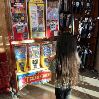H-E-B - Granddaughter eyeing on those candy and toy vending machine dispensers - A girl with long brown hair, wearing a light-colored top and dark leggings with heart patterns, is standing in front of a colorful vending machine labeled 'Texas Carnival.' The machine offers gumballs, toys, and tattoos, with various items visible inside. The store has wooden walls, and shelves on the right display umbrellas and other accessories. Sunlight casts shadows on the floor. - Grocery Near Me - Waco, Texas