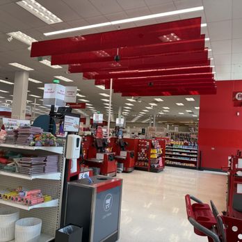 Target - The image shows the interior of a Target retail store, focusing on the checkout area with red checkout counters and digital screens. Shopping carts are visible on the right side, and shelves with various products are in the background. The ceiling has white panels with fluorescent lighting, and red decorative panels hang from the ceiling. Signage indicates self-checkout options and other store services. - Department Stores Near Me - Skokie, Illinois