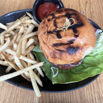 Wild Sage - A burger with a toasted bun, lettuce, and toppings is presented on a black plate. The burger is secured with a toothpick and has grill marks on the bun. Accompanying the burger are French fries and a small black container of ketchup. The plate is placed on a wooden table, and the image captures the meal from a top-down perspective. - Salad Near Me - Eagle, Colorado