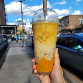 Heytea - Coconut Mango Boom - A person holds a large bubble tea with mango and coconut flavors, featuring a straw and a clear lid, against a city street background with buildings and cars. - Bubble Tea Near Me - Brooklyn, New York