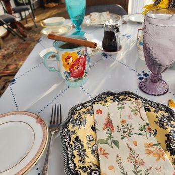 Everitt House - The table is set with a floral-patterned plate, a matching napkin, and a fork. There is a coffee mug with a painted floral design, containing a cinnamon stick, and a small pitcher of syrup. Colorful glassware, including a blue water glass and a purple goblet, are also on the table. The background shows a cozy, vintage-style room with wooden furniture and patterned rugs. - Bed & Breakfast Near Me - Hackettstown, New Jersey